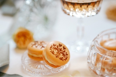 Zwei goldene Macarons mit filigranem Muster auf Glasschale, daneben Kristallglas und Blume auf elegant gedecktem Tisch.