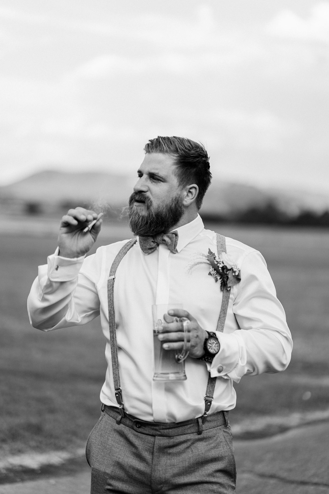 Ein Mann im Anzug mit Bart und Fliege raucht und hält ein Bierglas in der Hand, draußen auf einem Feld.