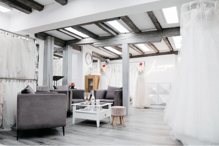 Heller Brautladen mit weißen Kleidern auf Ständern, grauen Sofas, kleinem Tisch und Holzbalkendecke.