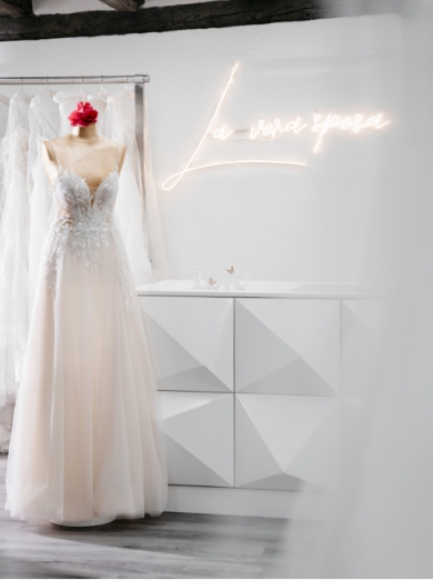 Schaufensterpuppe mit weißem Brautkleid und Blumenkranz, daneben ein leuchtendes La vera sposa-Schild auf weißer Wand.