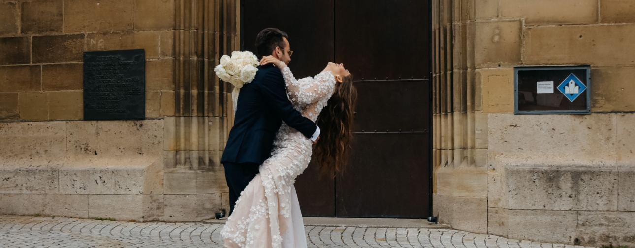 Ein Hochzeitspaar tanzt vor einer steinernen Wand; der Bräutigam hält die Braut, die Blumen trägt.