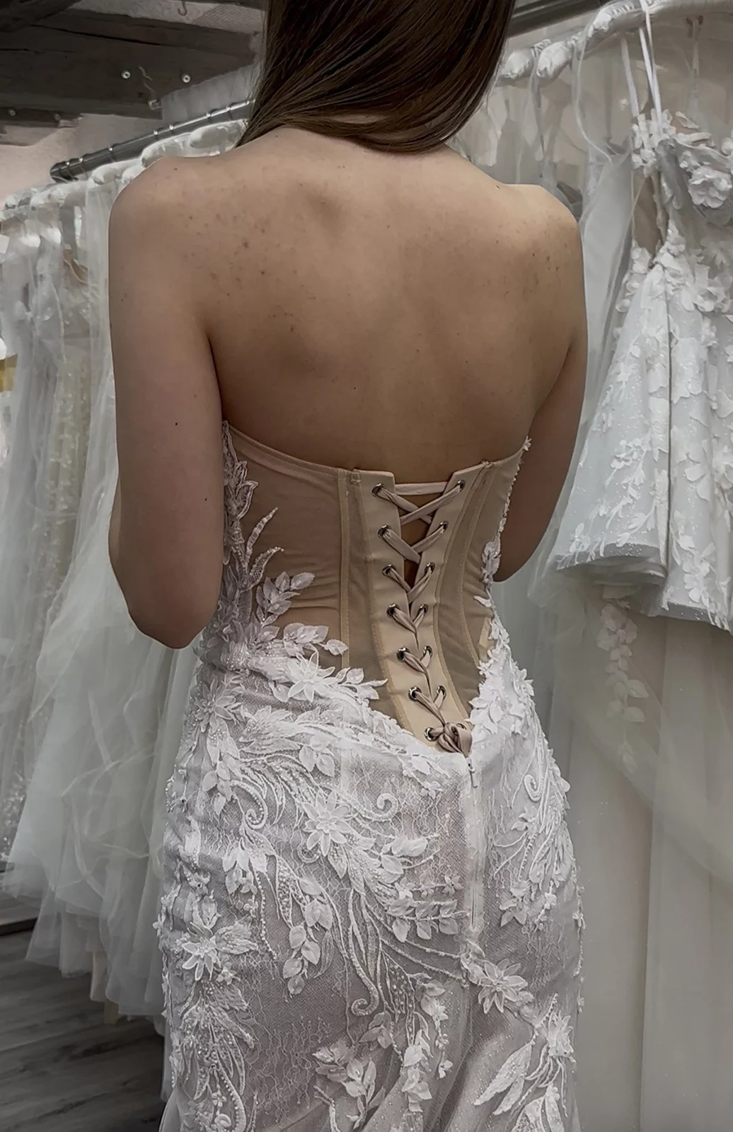 Frau in Hochzeitskleid mit Spitzendetails und Schnürung schaut auf Kleiderstange in einem Brautmodengeschäft.