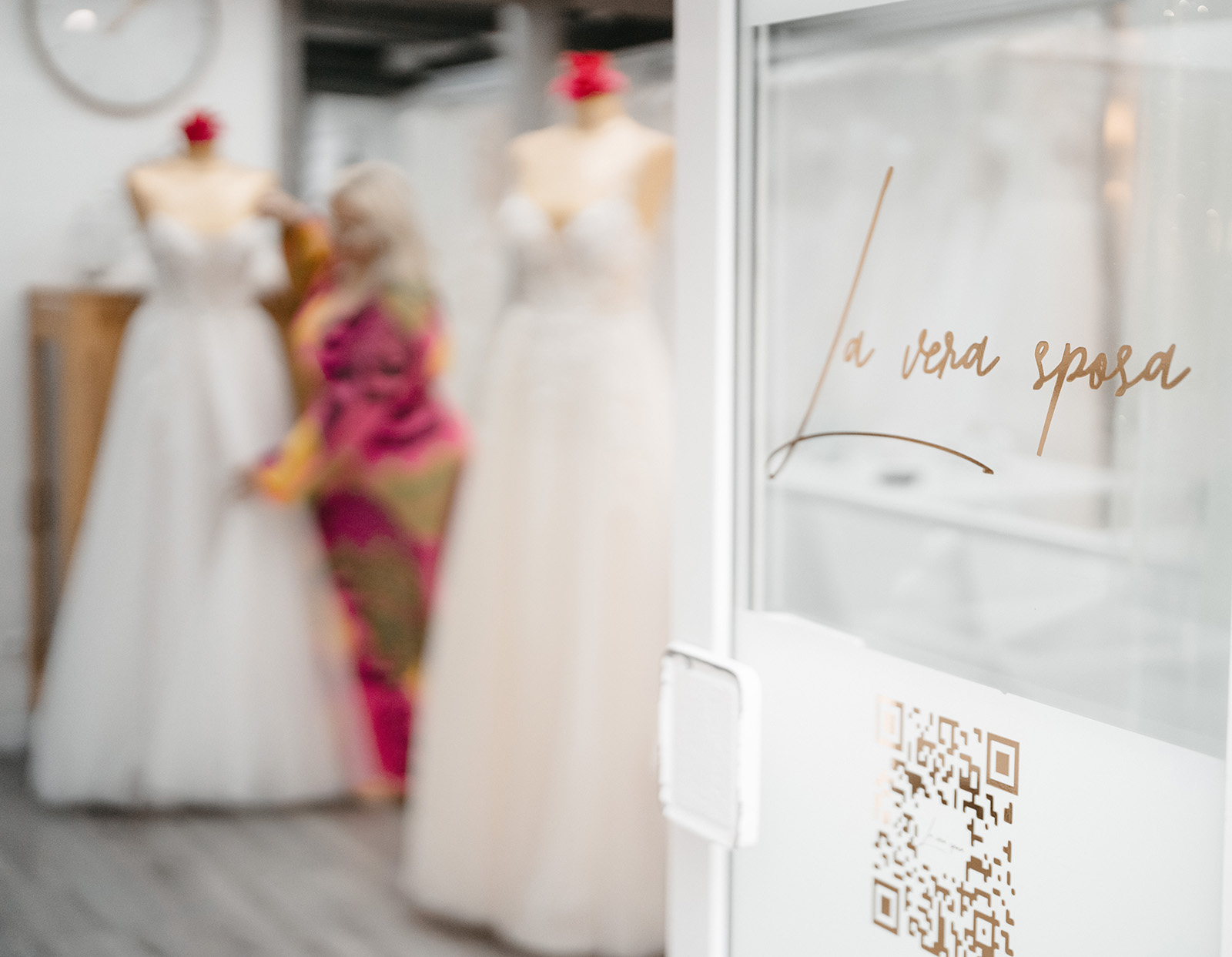 Schaufenster mit QR-Code, Aufschrift La vera sposa. Unscharfe Ansicht von Hochzeitskleidern und Person im Hintergrund.