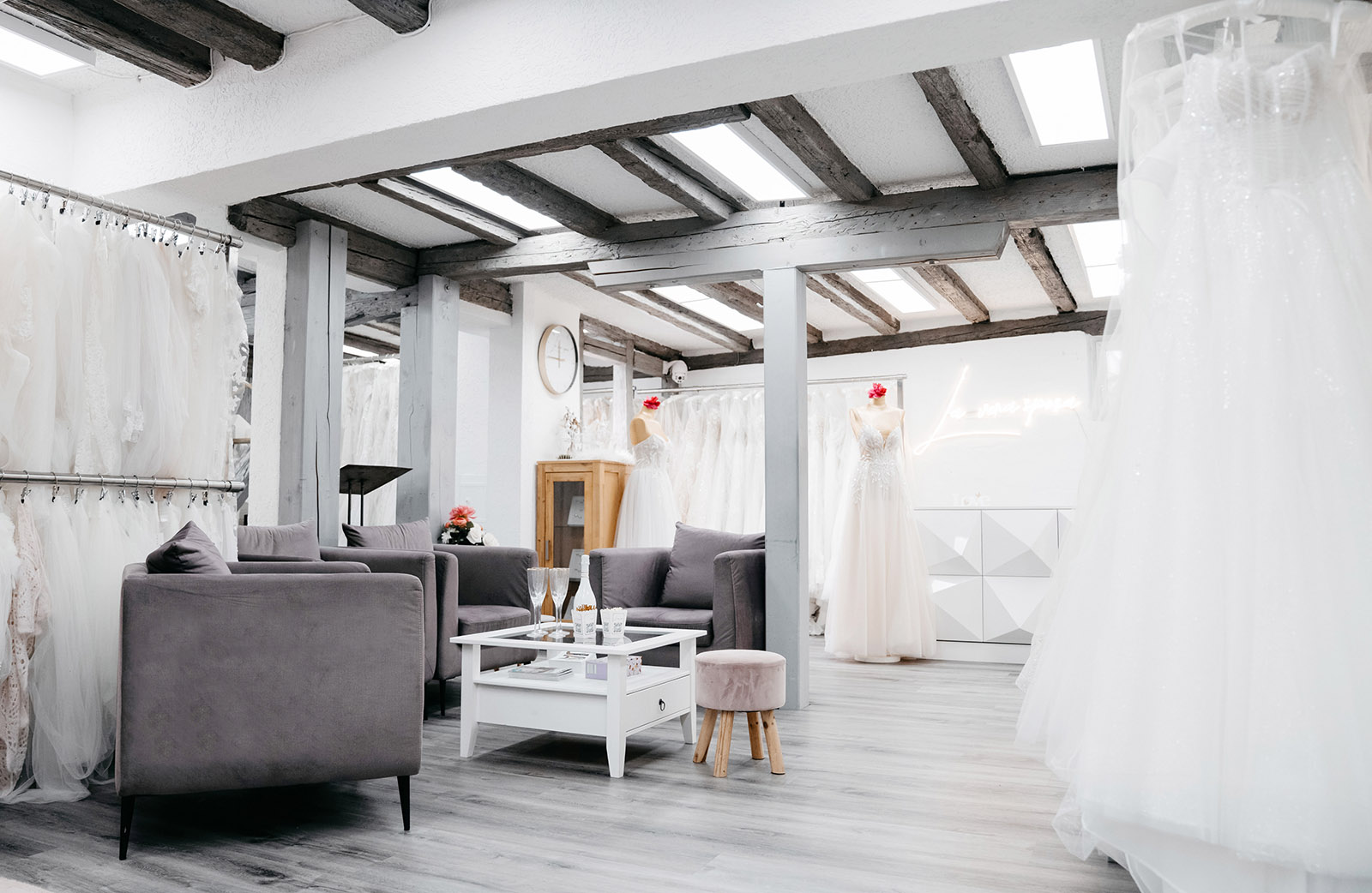 Brautmodengeschäft mit weißen Kleidern, lila Sofas, Couchtisch, Mannequins in Brautkleidern, helle Holzdeckenbalken.