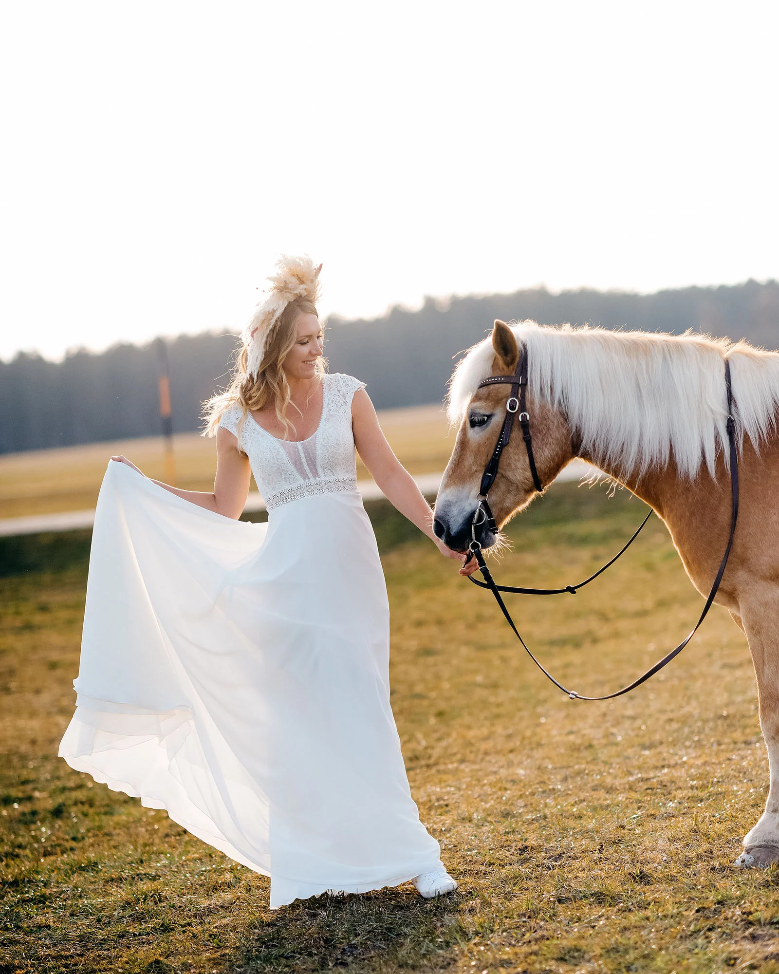 Frau in weißem Kleid führt ein braunes Pferd auf einer Wiese bei Sonnenuntergang. Sie lächelt und hält das Kleid hoch.