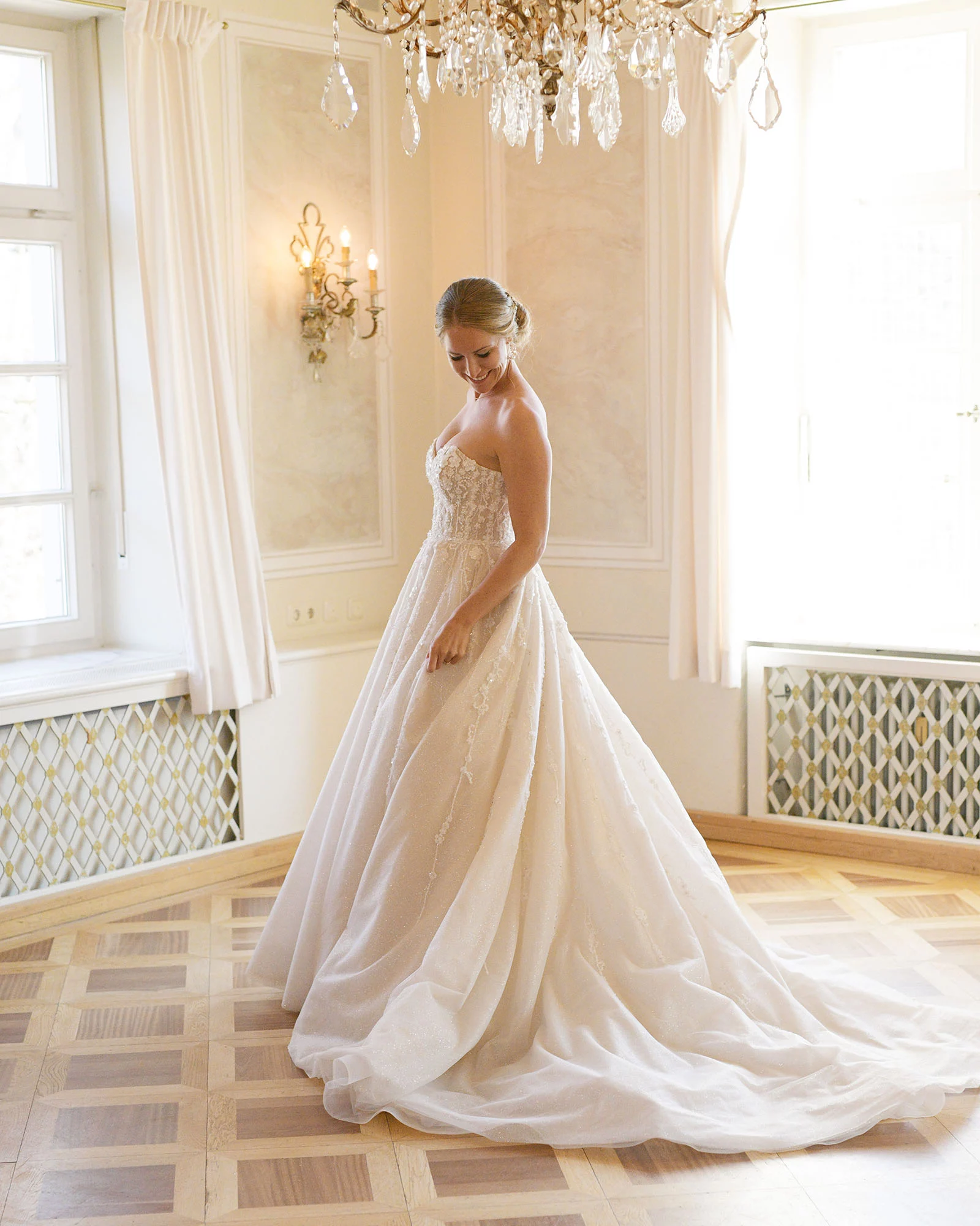 Braut in einem weißen, schulterfreien Kleid steht in einem eleganten Raum mit Kronleuchter und blickt nach unten.