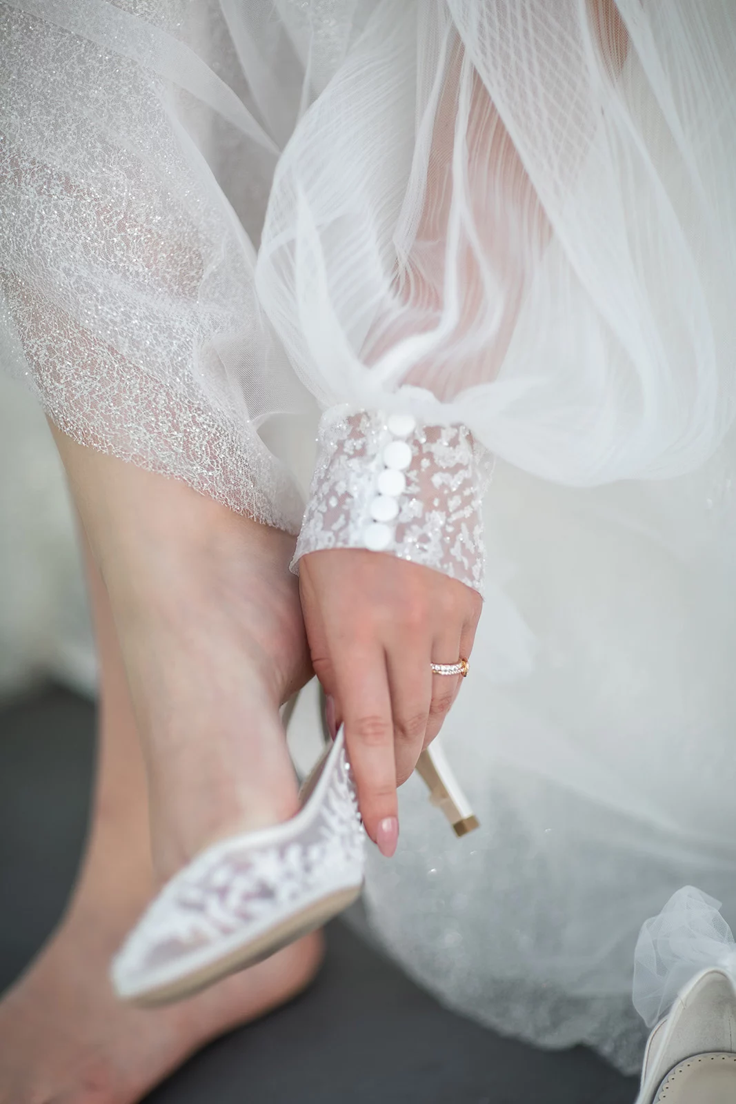 Hand hält eleganten weißen Schuh, Teil eines Spitzenkleides sichtbar. Person trägt einen Ring.