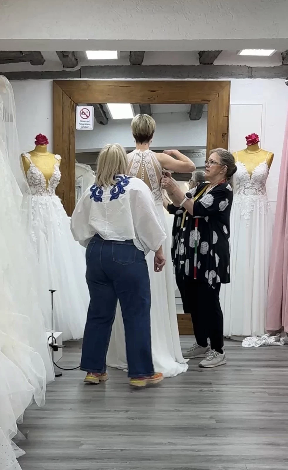 Zwei Frauen helfen einer Person beim Anpassen eines Brautkleids in einem Geschäft mit Spiegel und Schaufensterpuppen.