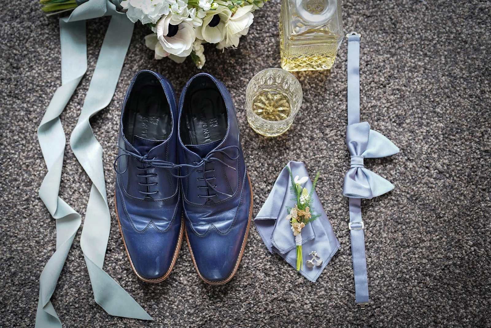 Blaue Herrenschuhe, Fliege, Einstecktuch mit Blume, Glas und Karaffe mit Whiskey auf grauem Teppich arrangiert.