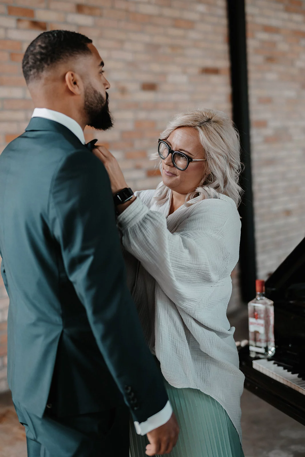 la-vera-sposa_moderner-Braeutigam_blogbeitrag_02 Mann im Anzug und Frau mit Brille stehen in modernem Innenraum, sie richtet seine Krawatte; Klavier und Wodkaflasche im Hintergrund.